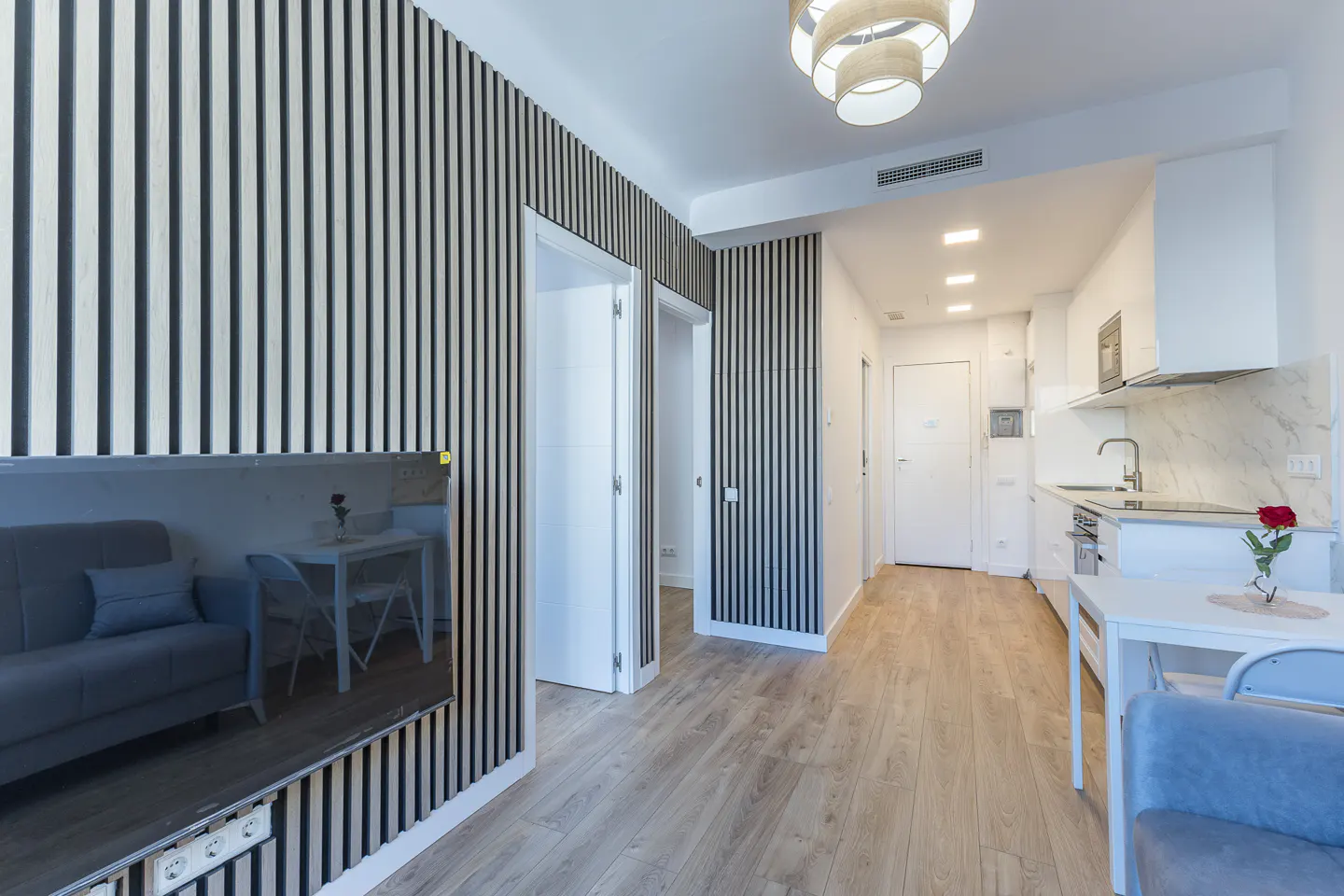 Modern apartment interior with wood-slat accent walls, a grey sofa, a small dining area, and a white kitchen.