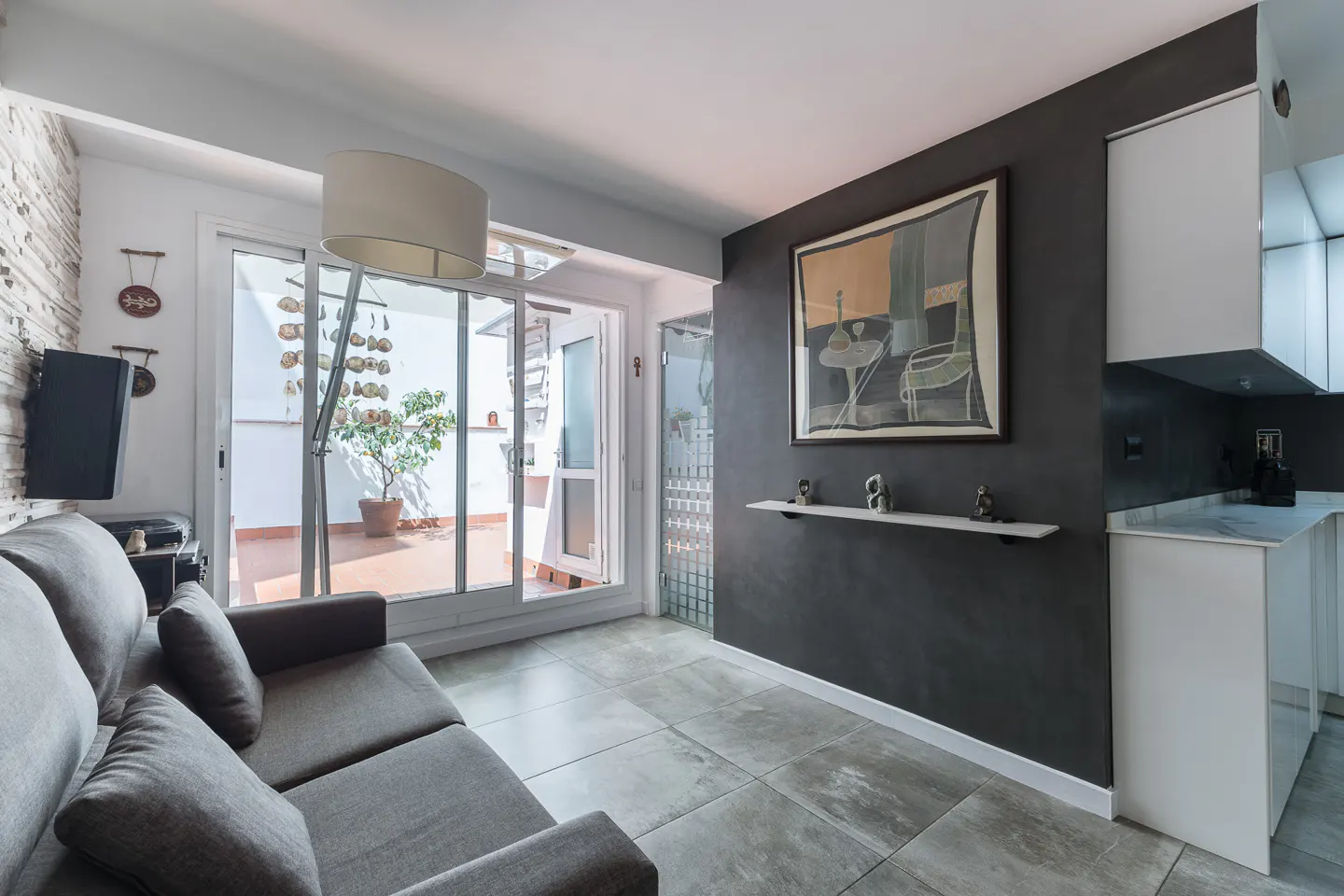 Living room with gray sofa, TV, and sliding glass doors to a patio with a potted lemon tree. A dark gray wall features art and a shelf.