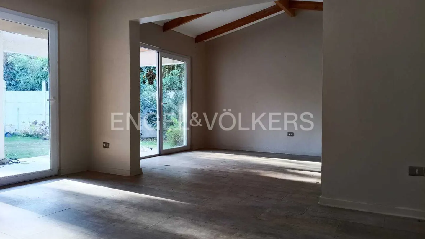 Empty room with light-colored walls, tile floors, wooden ceiling beams, and large glass doors leading to a garden.