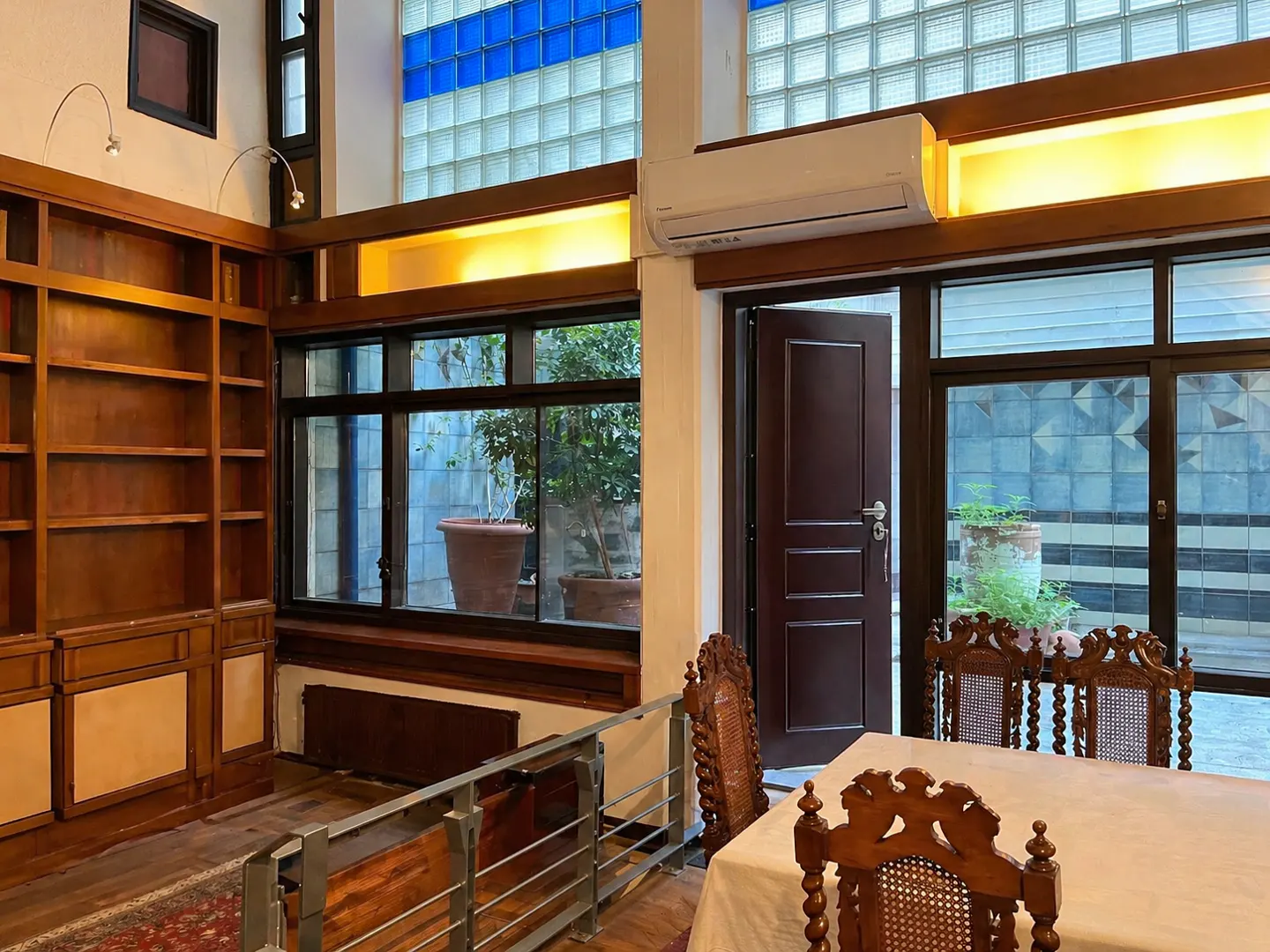 Interior view of a dining room with a wooden table, chairs, and a bookshelf. A window overlooks a courtyard with plants.