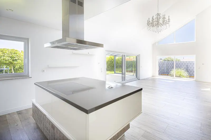 Modern white kitchen with a central island, stainless steel hood, and bright open-plan living area with a chandelier.