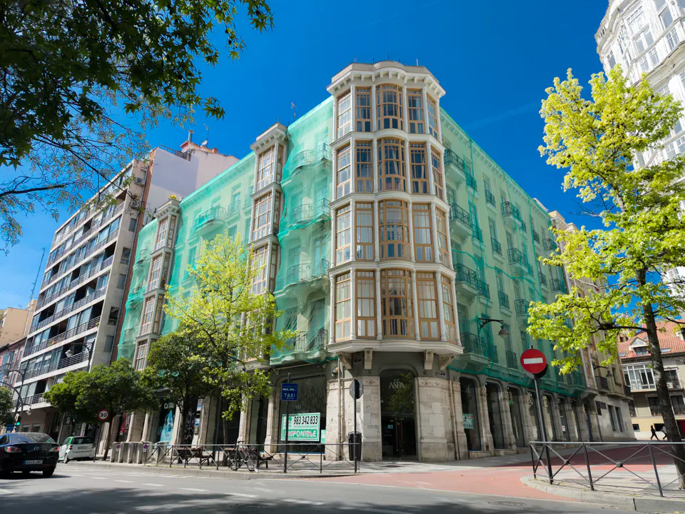 Street view of a tall building covered in green netting for construction, under a bright blue sky.