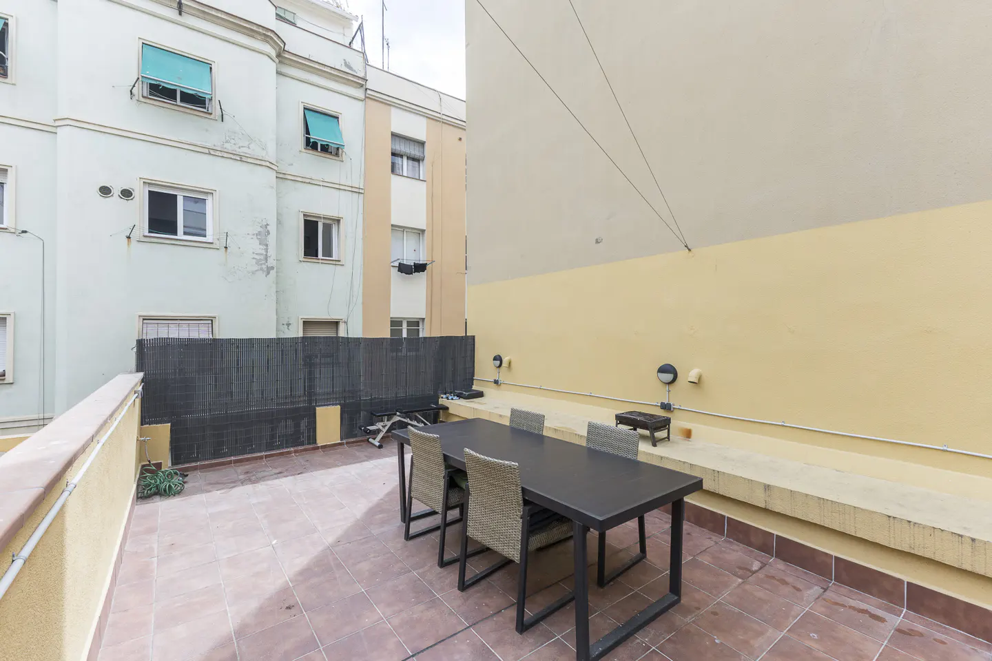 Outdoor patio with a black table and four chairs on a red tiled floor. Buildings surround the patio.