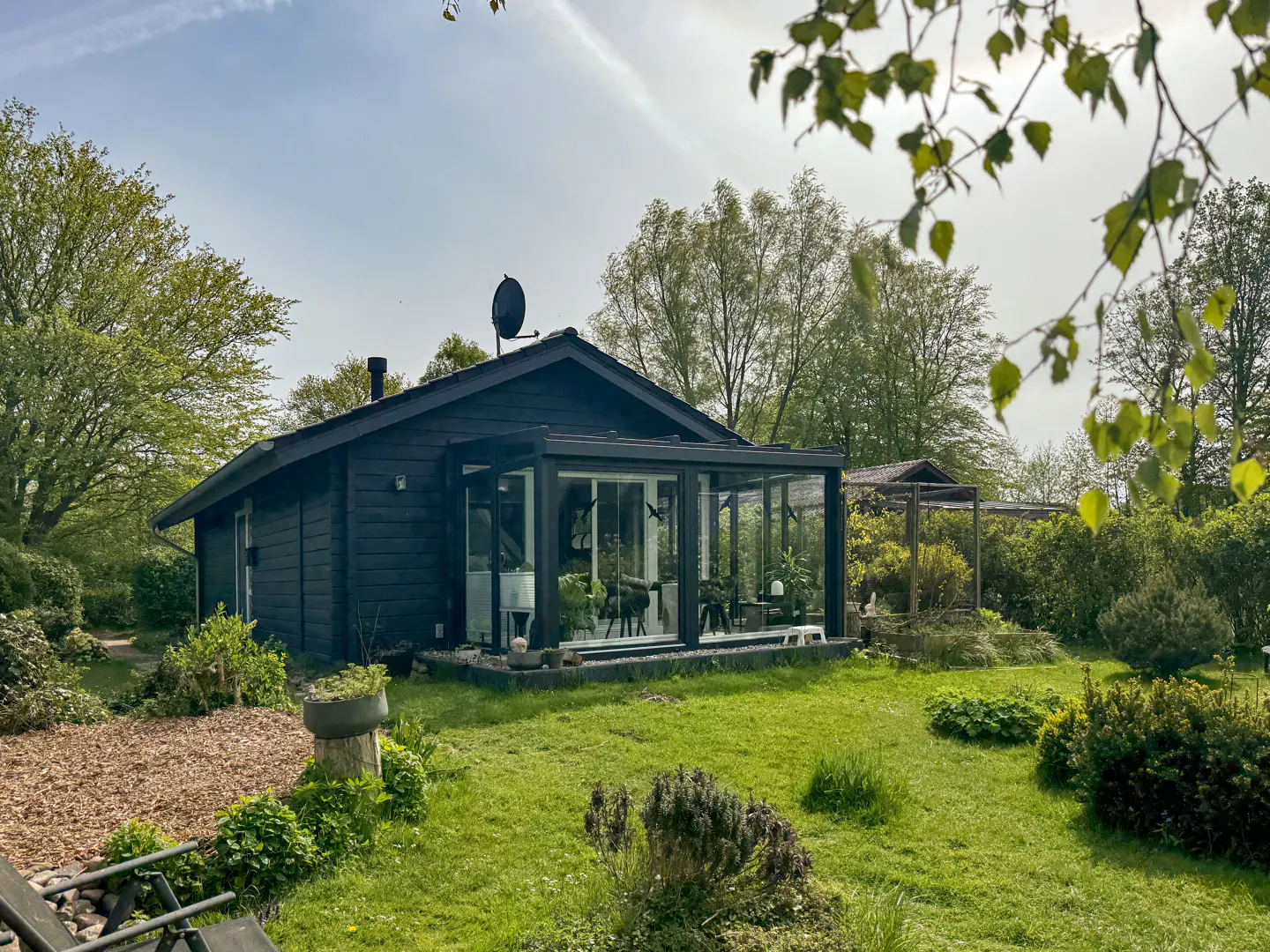 A black wooden cabin with a glass-enclosed sunroom sits in a lush green garden surrounded by trees on a sunny day.