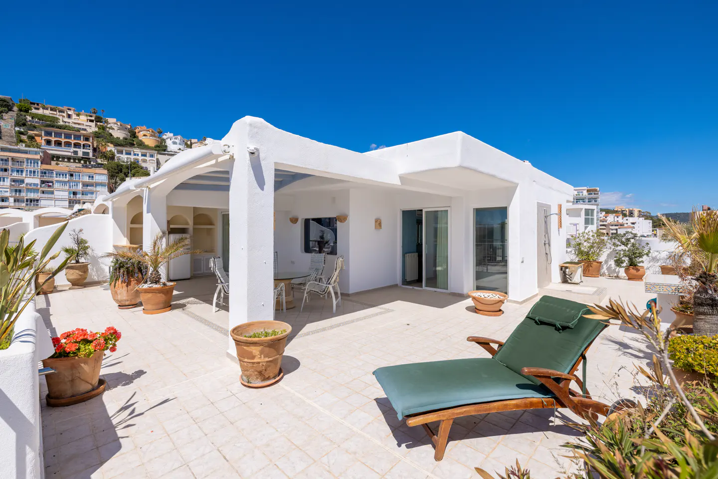 Bright rooftop patio with white walls, tile floor, and blue sky. Green lounge chair, potted plants, and outdoor dining set.