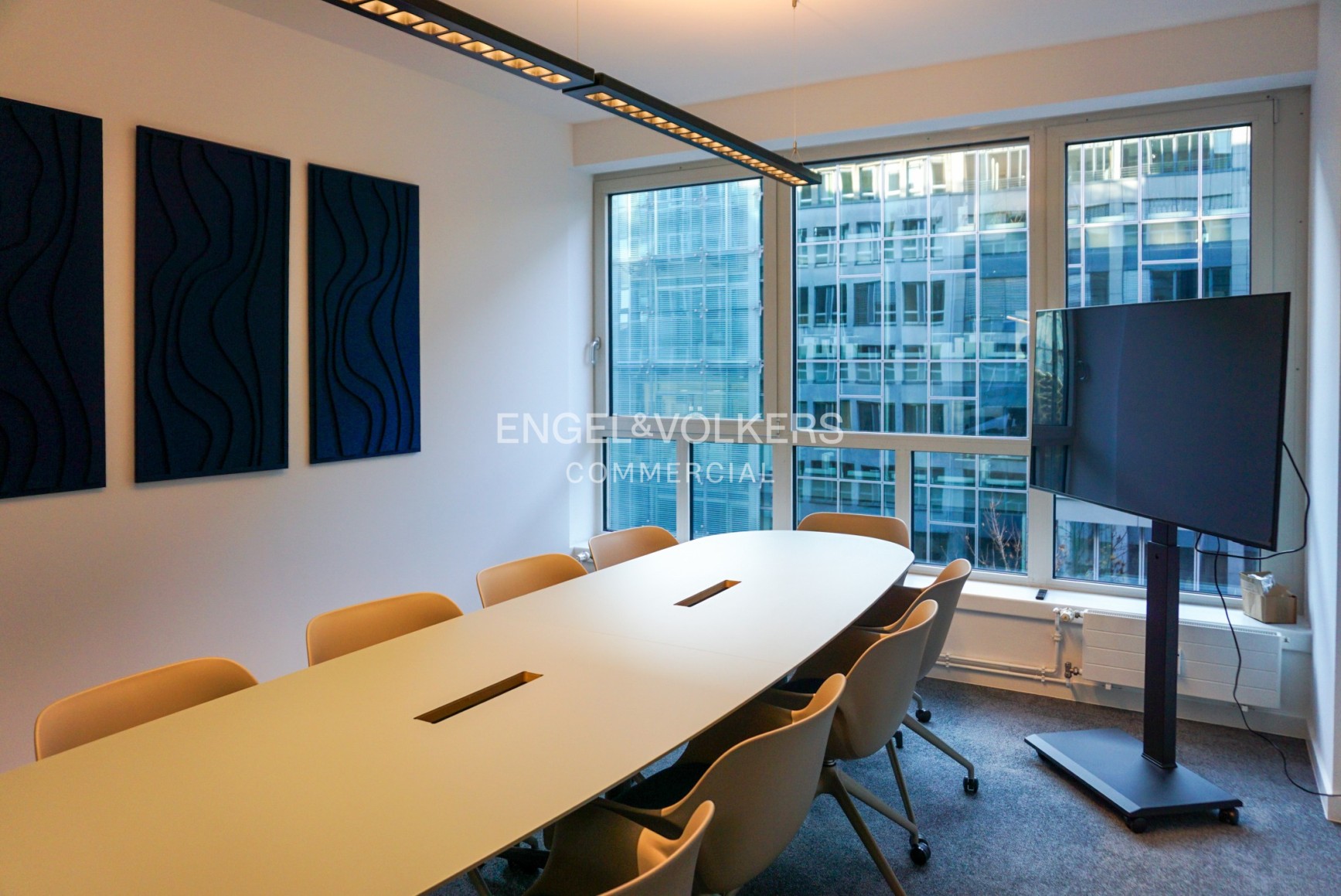 Modern conference room with a long white table, tan chairs, wall-mounted acoustic panels, and a large TV on a stand.