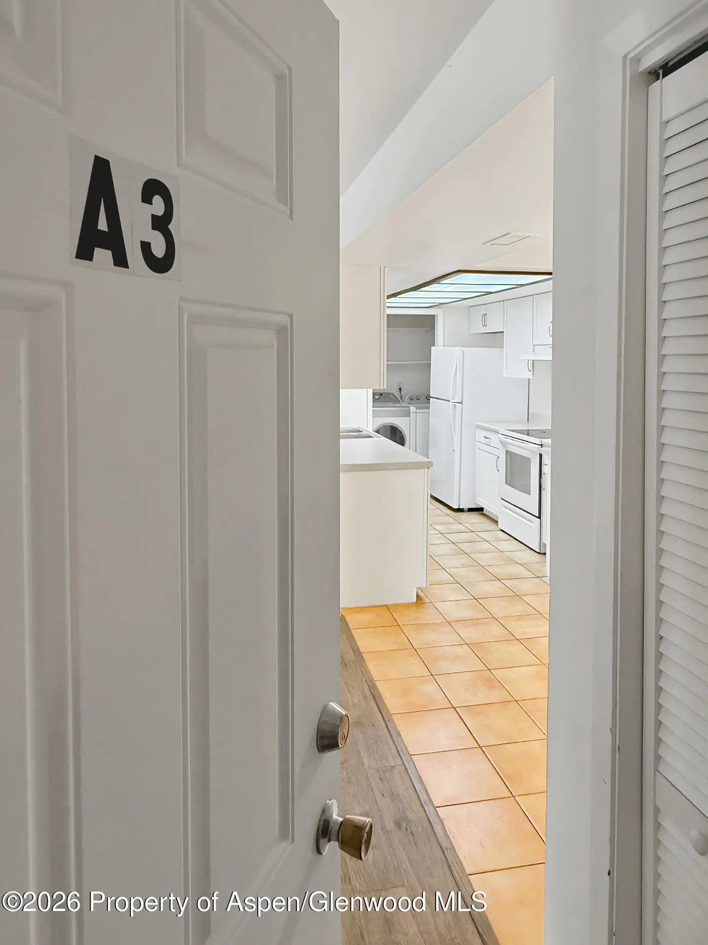 Open white door with "A3" on it, revealing a kitchen with white appliances and tan tile flooring.