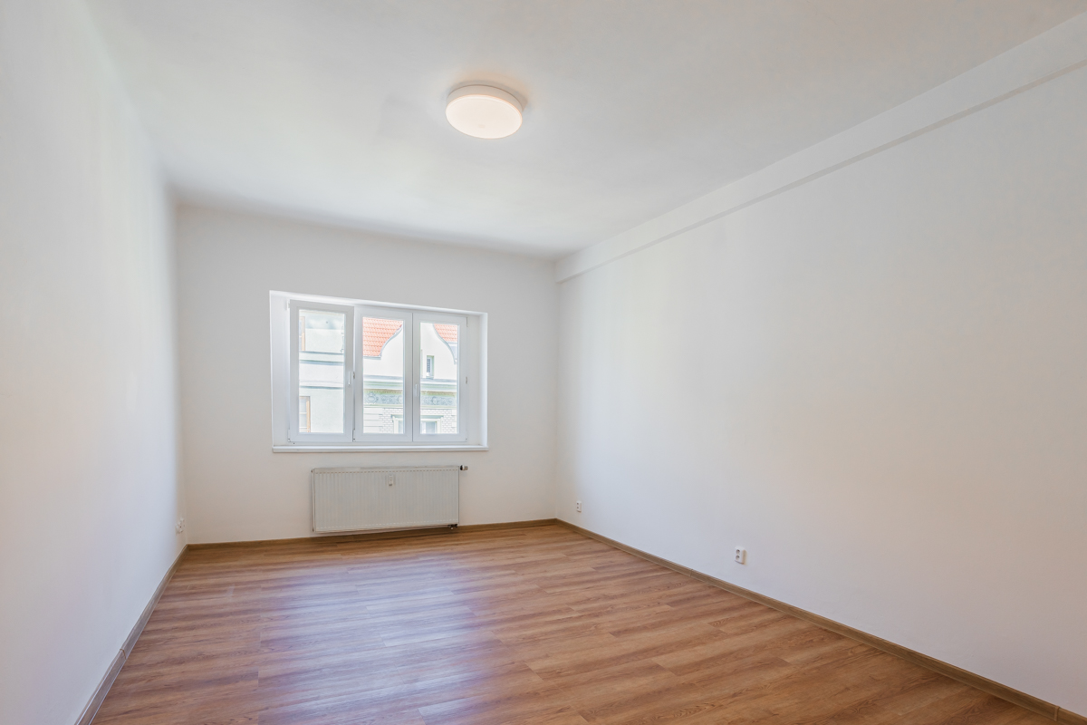 Empty room with white walls, light wood laminate flooring, a central ceiling light, and a window with a radiator below.
