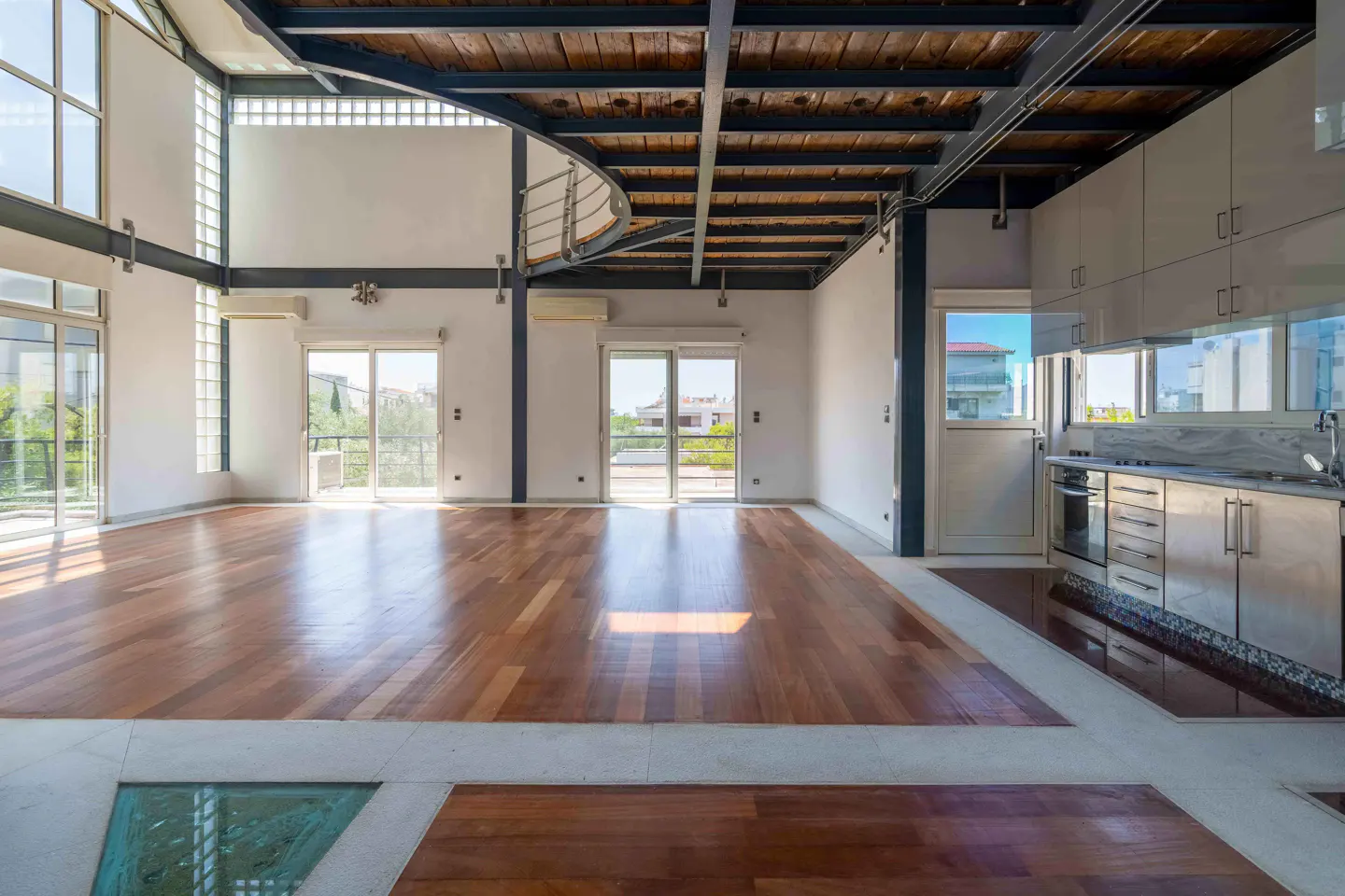 Bright, modern loft with hardwood floors, white walls, and exposed ceiling beams. Kitchen with stainless steel appliances visible. Balcony doors open to greenery.