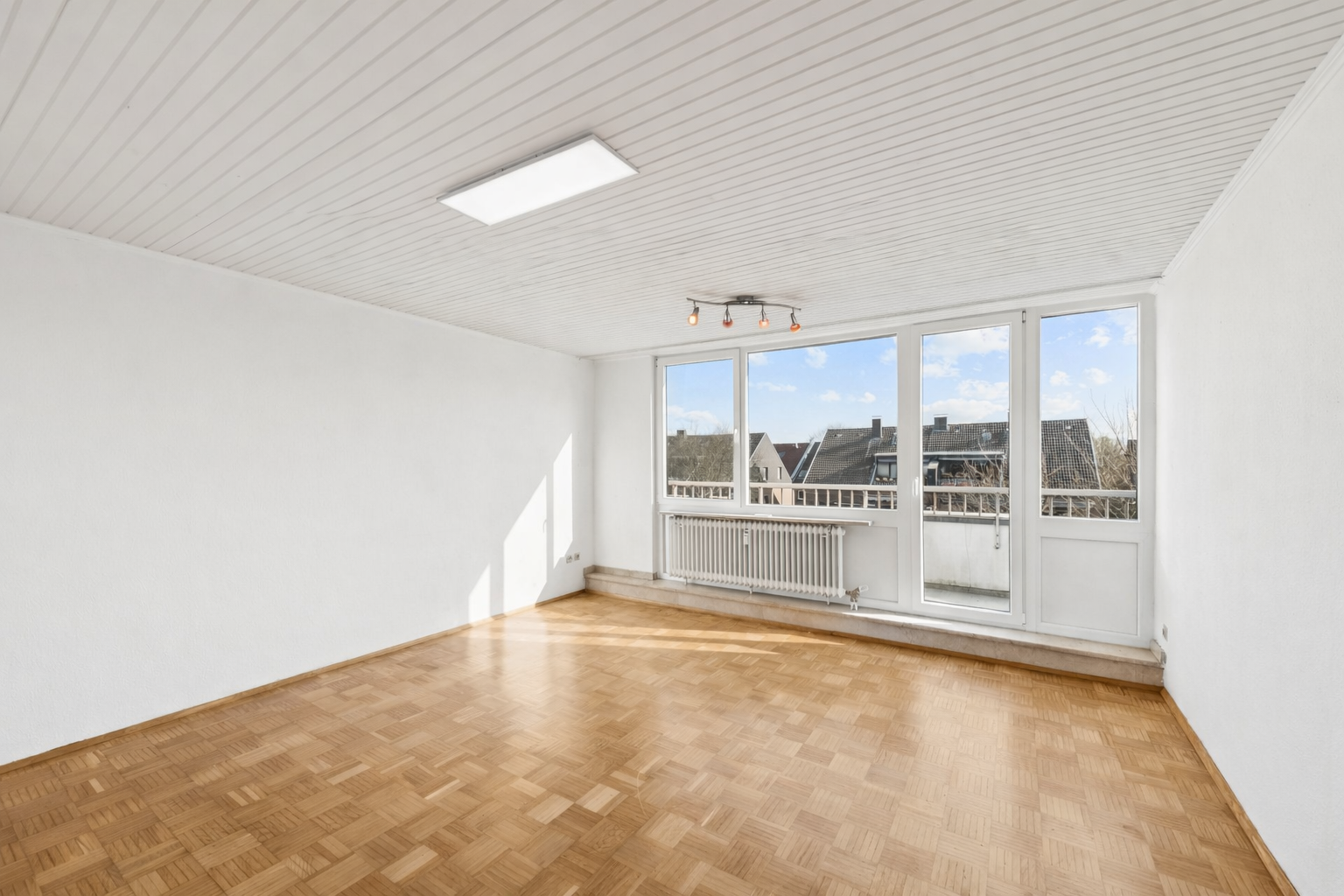 Empty room with white walls, wood parquet flooring, a paneled ceiling, and large windows leading to a balcony.
