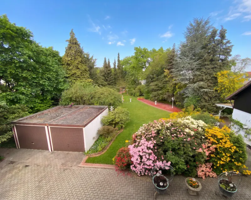 View of a lush garden with colorful flowers, green lawn, trees, and a two-car garage under a blue sky.