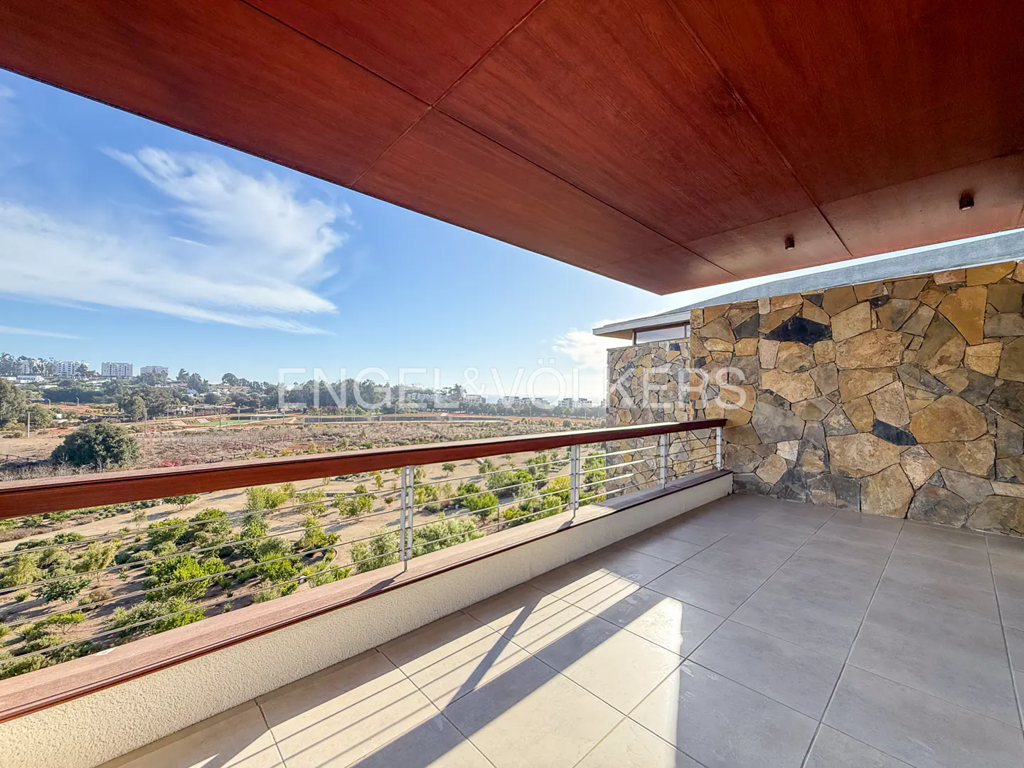 Balcony view with stone wall, wood ceiling, and metal railing overlooking a green landscape and distant city skyline.