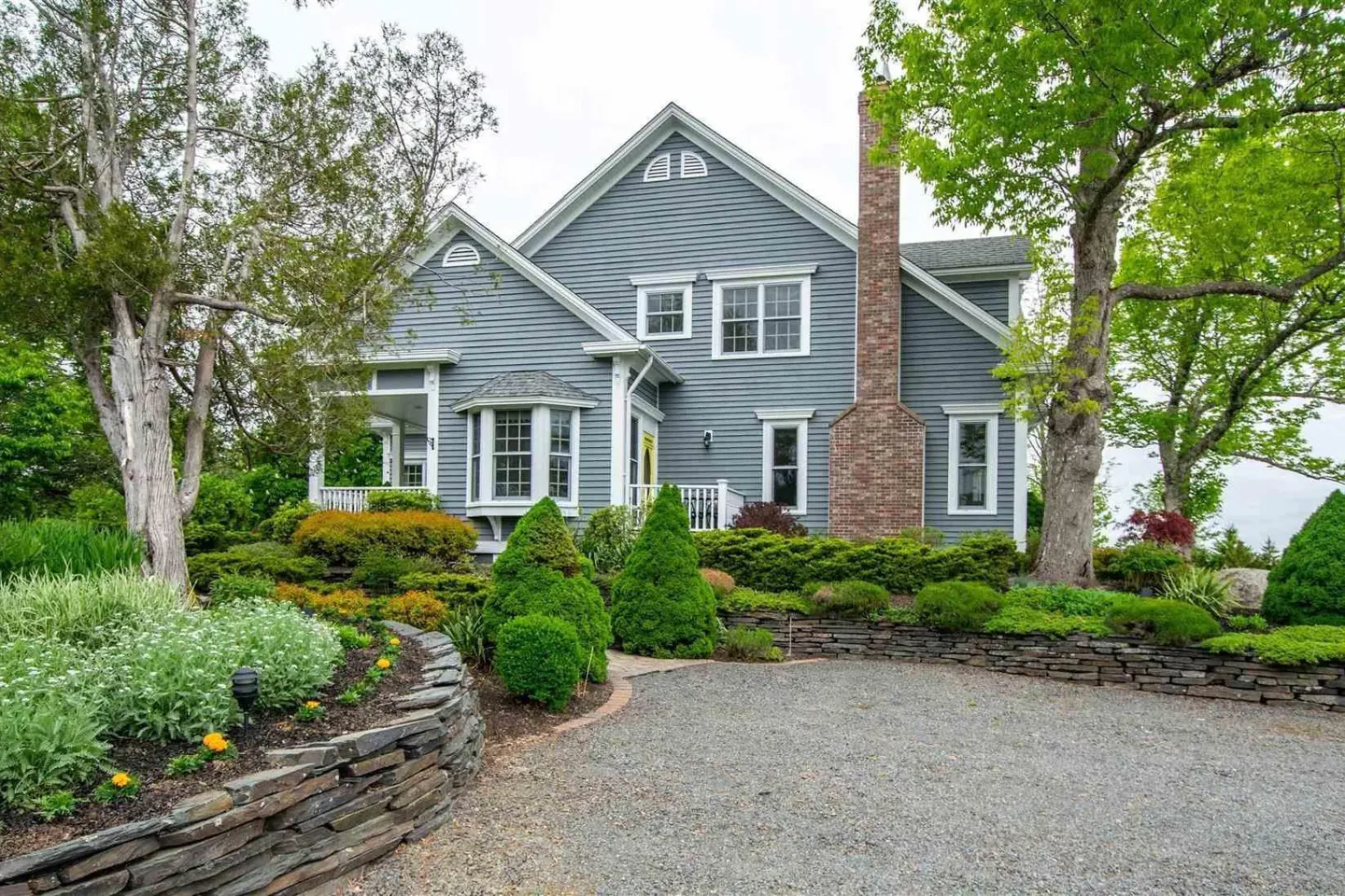 Two-story gray house with white trim, brick chimney, and manicured landscaping. Gravel driveway.