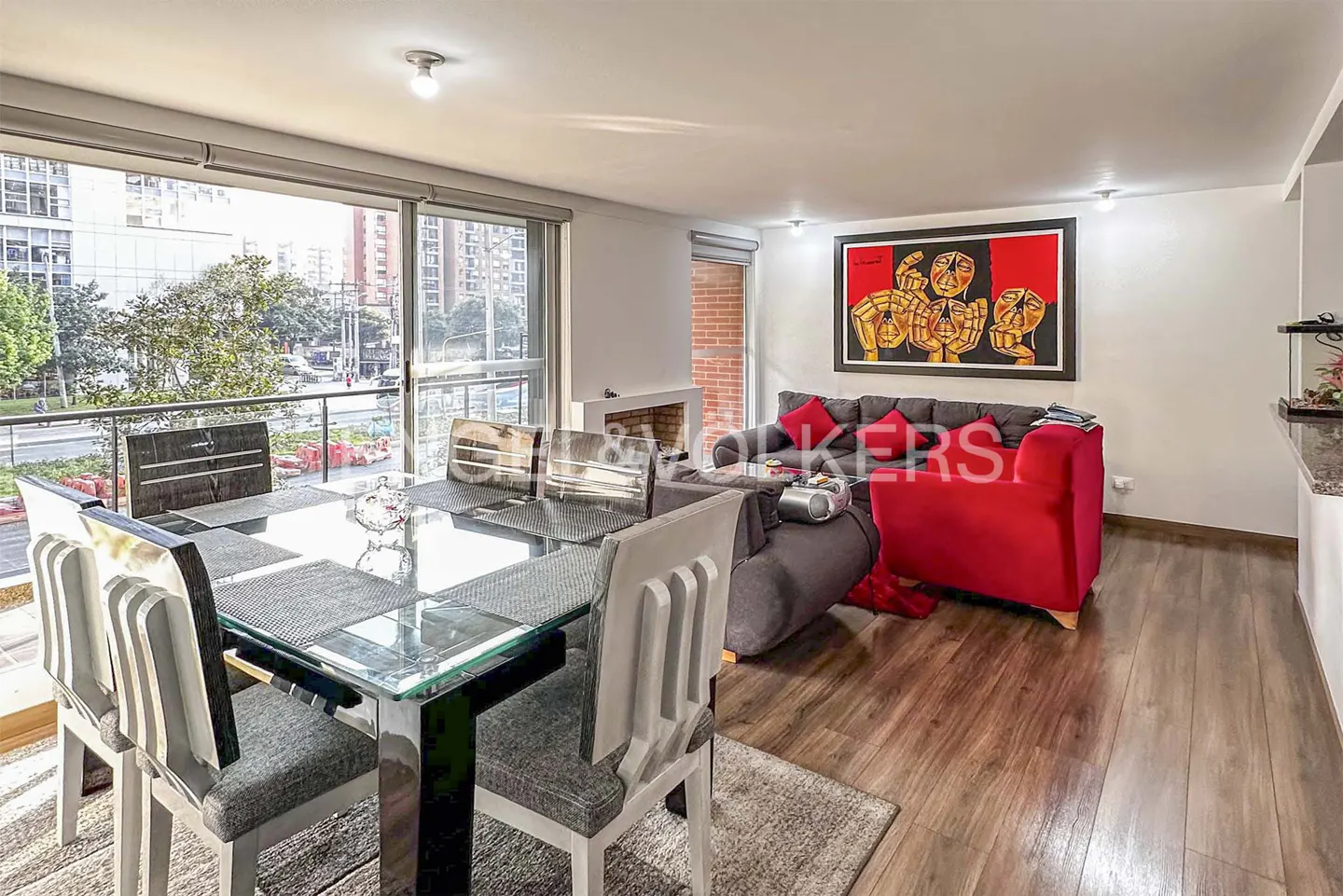Modern dining area with glass table and grey chairs, opening to a living room with a red sofa and abstract wall art.