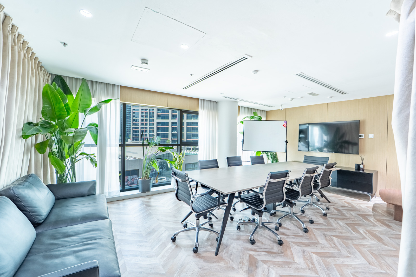 Modern conference room with a long table, black office chairs, a sofa, a whiteboard, and a large window with city views.