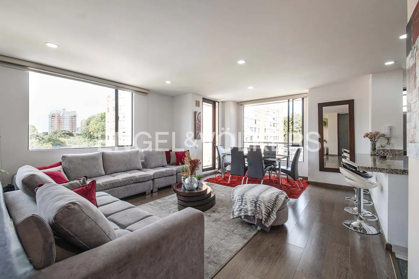 Bright, modern living room with a gray sectional sofa, red pillows, dining table, and breakfast bar. Large windows offer city views.