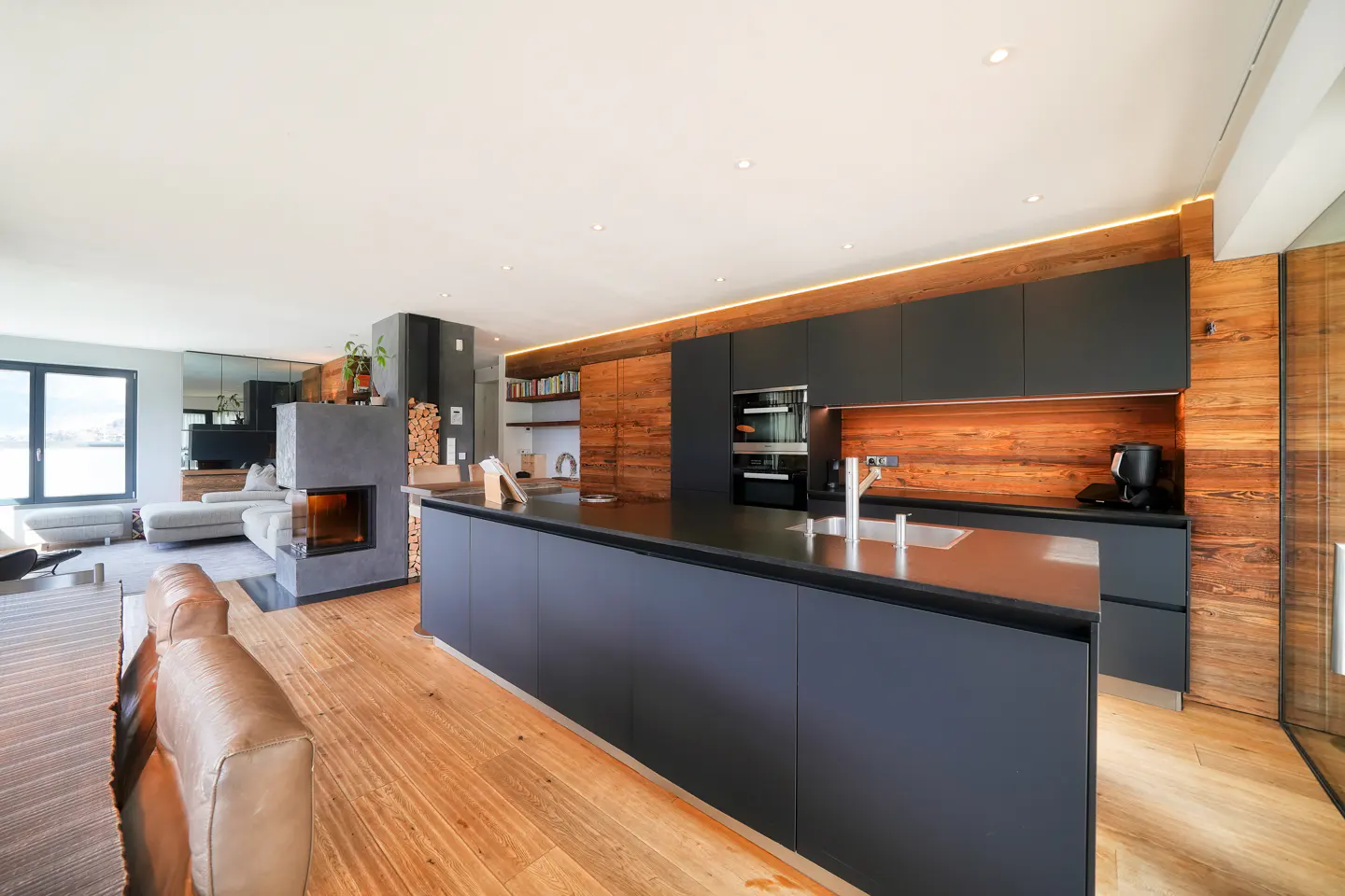 Open-concept living space with a black kitchen island, wood cabinets, and a fireplace. A tan leather couch is in the foreground.