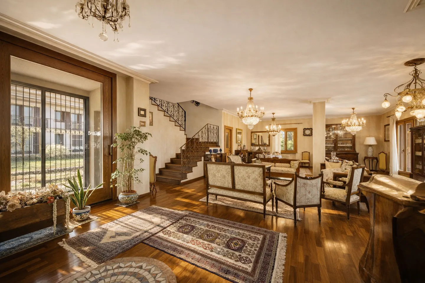 A spacious living room with hardwood floors, ornate rugs, and antique furniture. Chandeliers hang from the ceiling. A staircase leads to an upper level.