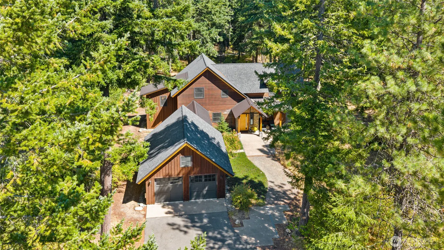 Aerial view of a brown wood-sided home with a dark roof, two-car garage, and yellow front door nestled in a pine forest.