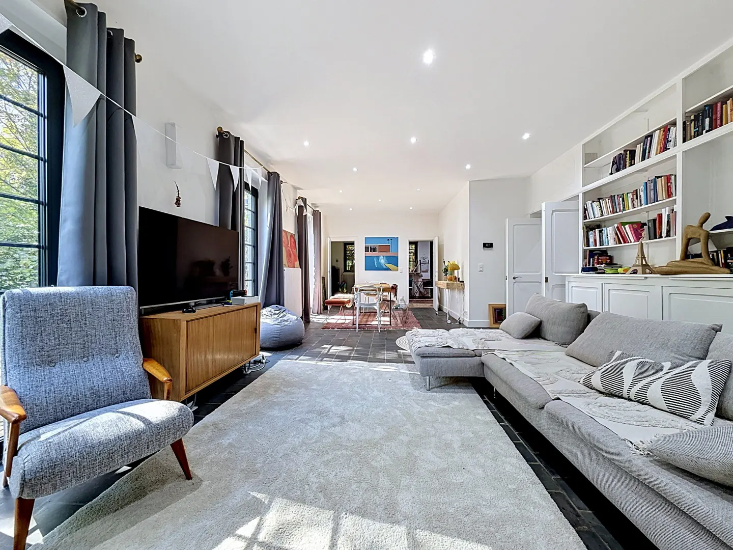 Bright living room with a gray sofa, chair, and rug. A TV sits on a wooden cabinet, and a bookshelf is built into the wall.