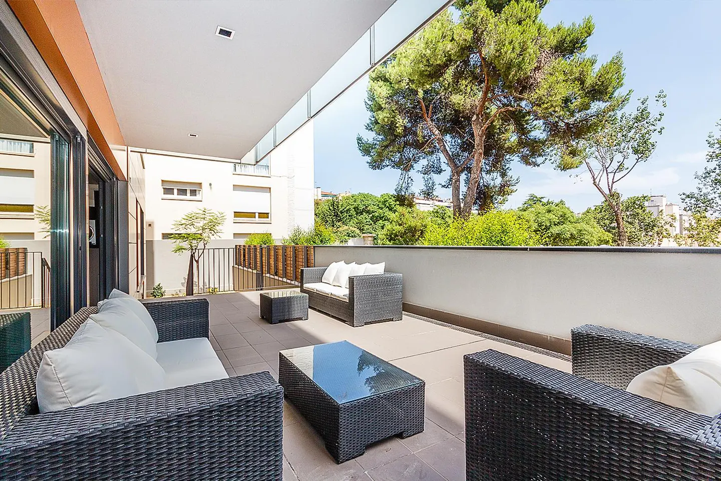 Outdoor patio with black wicker furniture, white cushions, and glass tables. Trees and buildings are visible in the background.