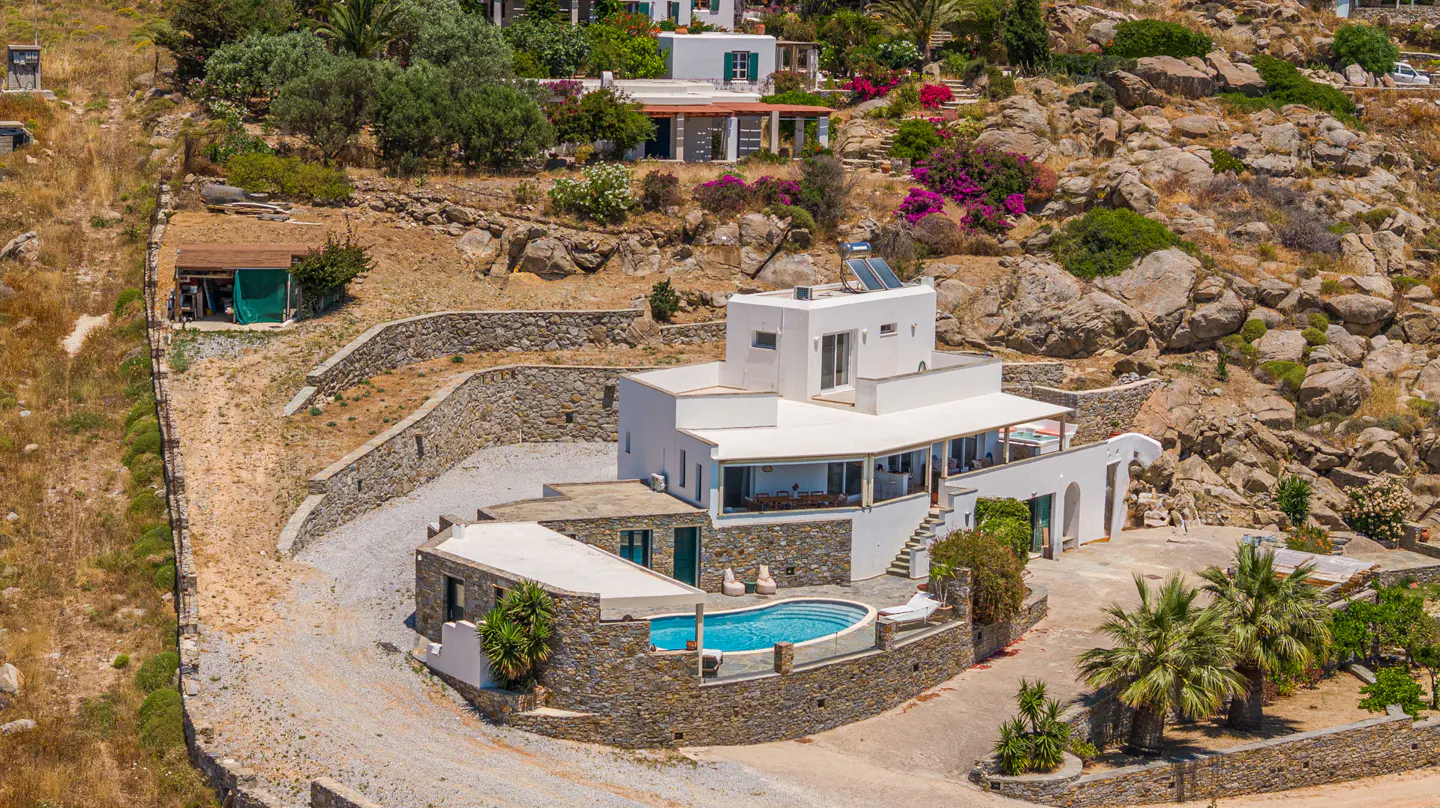 Aerial view of a white villa with a stone base and private pool, nestled into a rocky, sun-drenched hillside in Greece.
