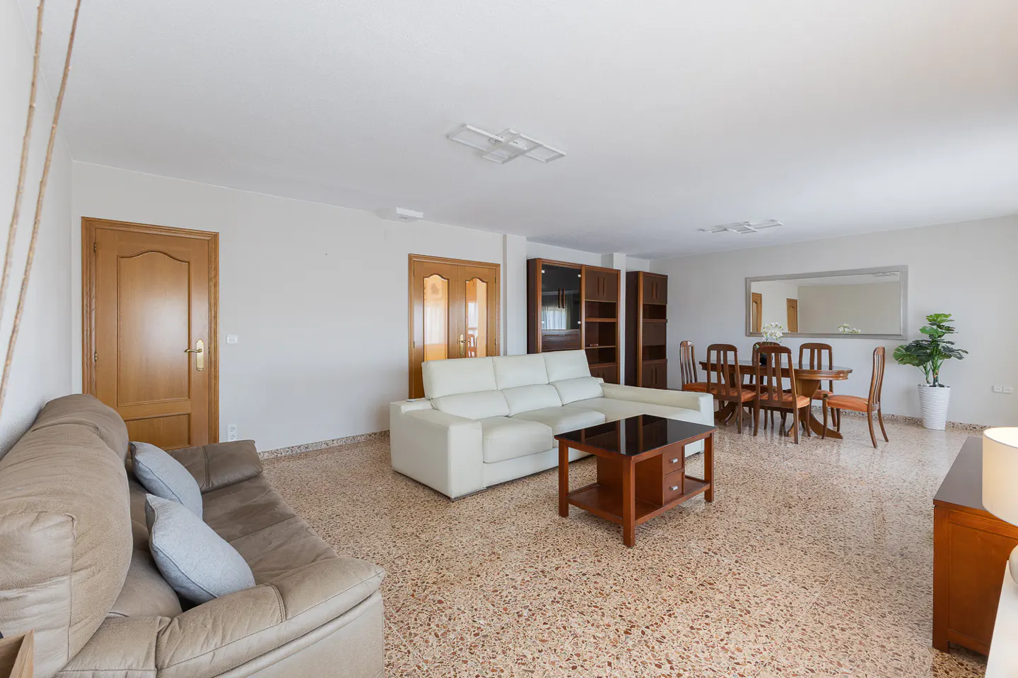 Bright living room with a brown sofa, white sectional, wood table, and dining set on a speckled floor.