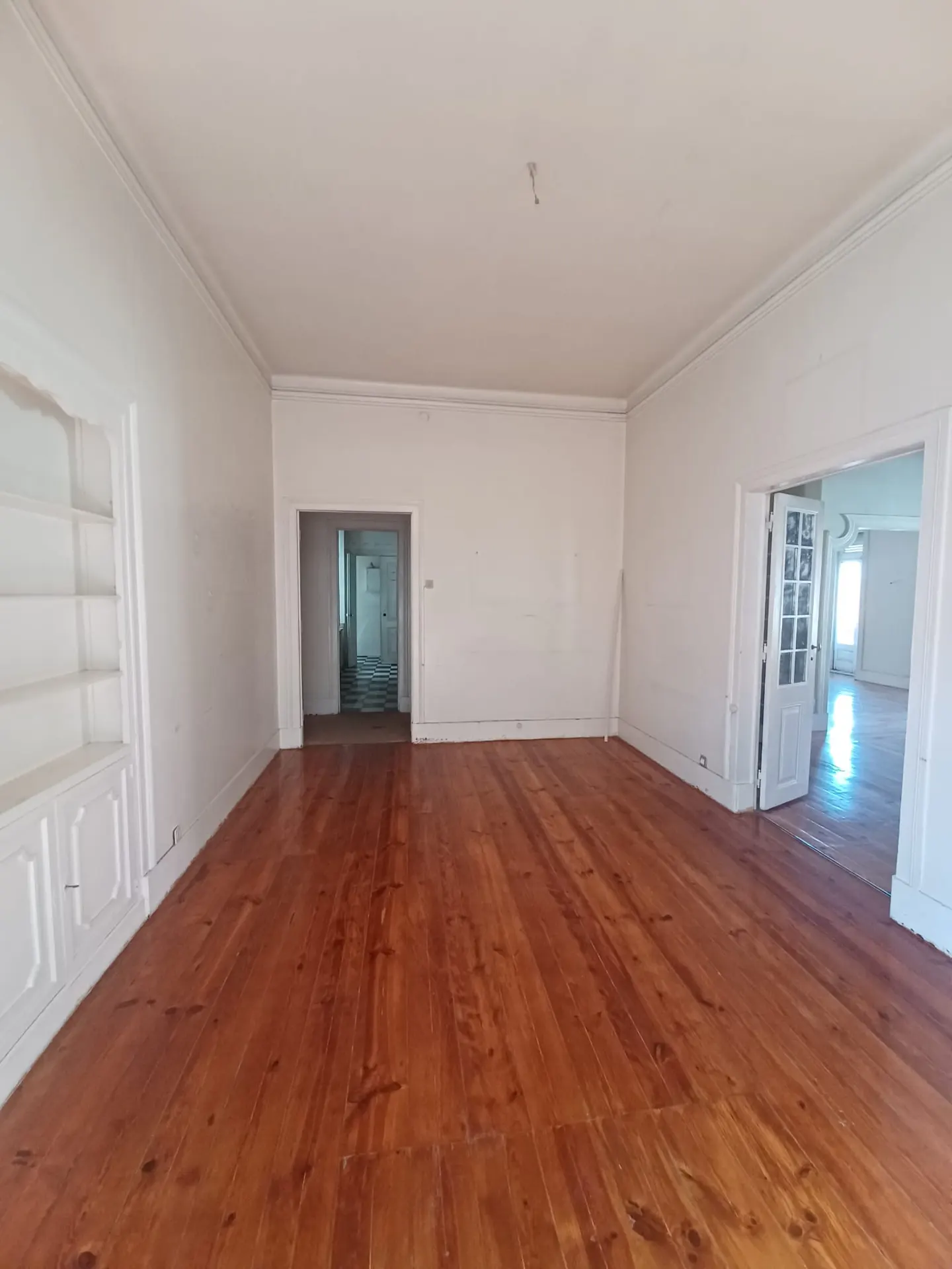 Empty room with hardwood floors, white walls, and doorways leading to other rooms. A built-in shelf is on the left.