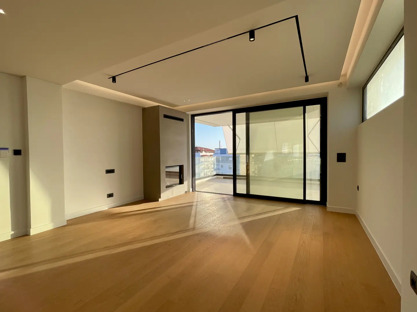 Bright, empty living room with hardwood floors, white walls, and a modern fireplace. Sliding glass doors lead to a balcony with a city view.
