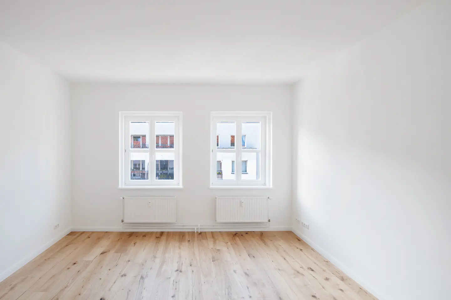 Bright, empty room with hardwood floors, white walls, and two white-framed windows. Radiators are under each window.