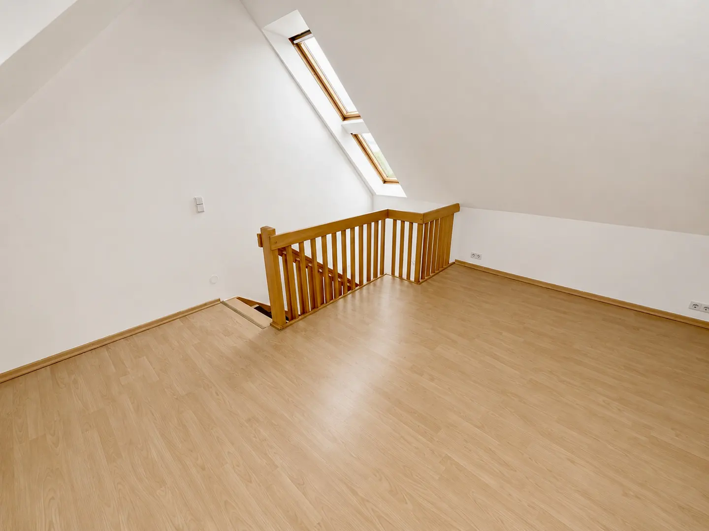 Attic room with light wood floors, white walls, and a wooden railing. Two skylights are visible in the sloped ceiling.