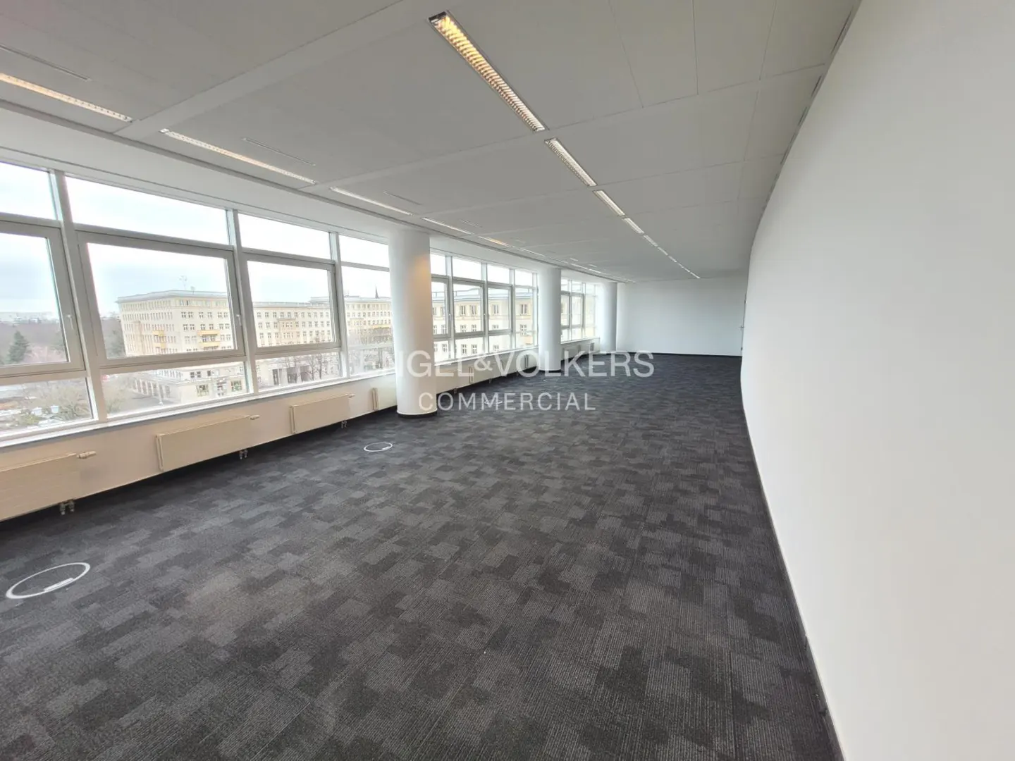Bright, empty office space with gray carpet, white walls, and large windows overlooking a city. "Engel & Volkers Commercial" logo visible.