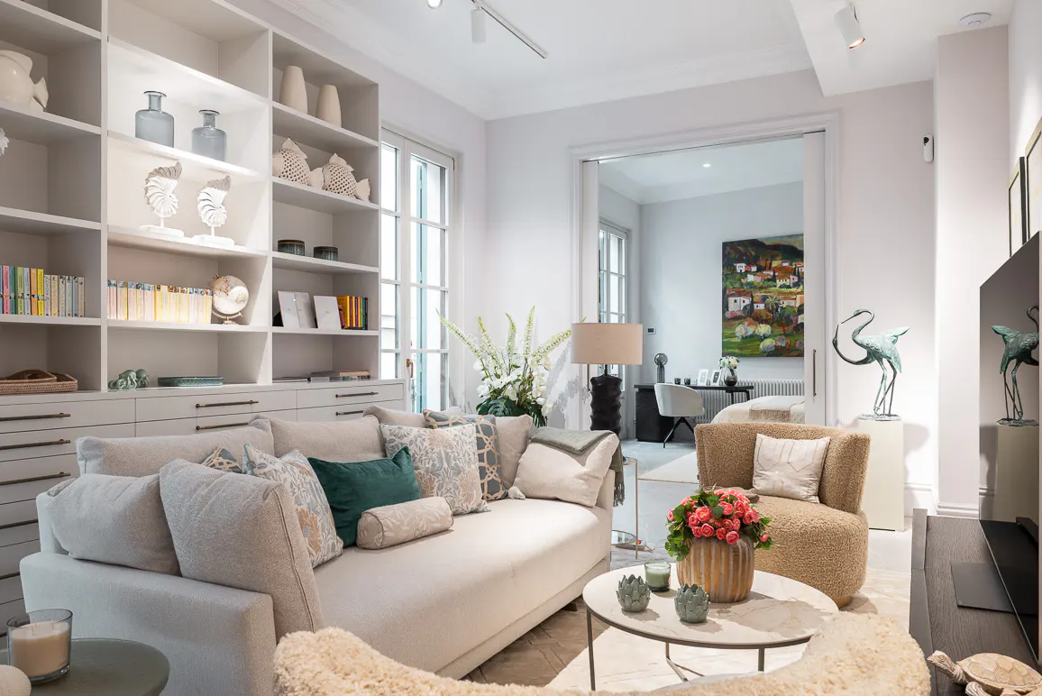 Bright living room with a white sofa, patterned pillows, and a built-in bookshelf filled with books and decorative items. A round coffee table sits in front.