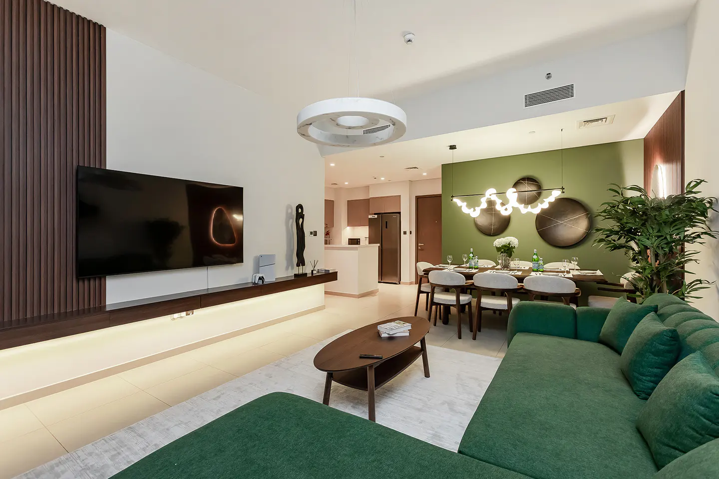 A modern living room with a green sofa, TV, and dining area. The walls are white and green, with wood accents. A round light fixture hangs above.
