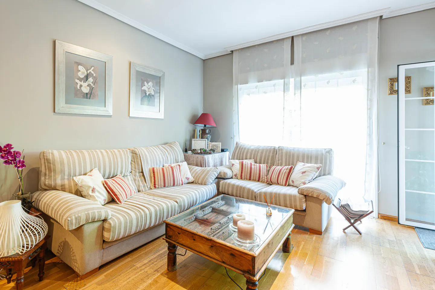 Living room with striped sofas, red pillows, a wooden coffee table, and hardwood floors. Two framed flower prints hang on the gray wall.