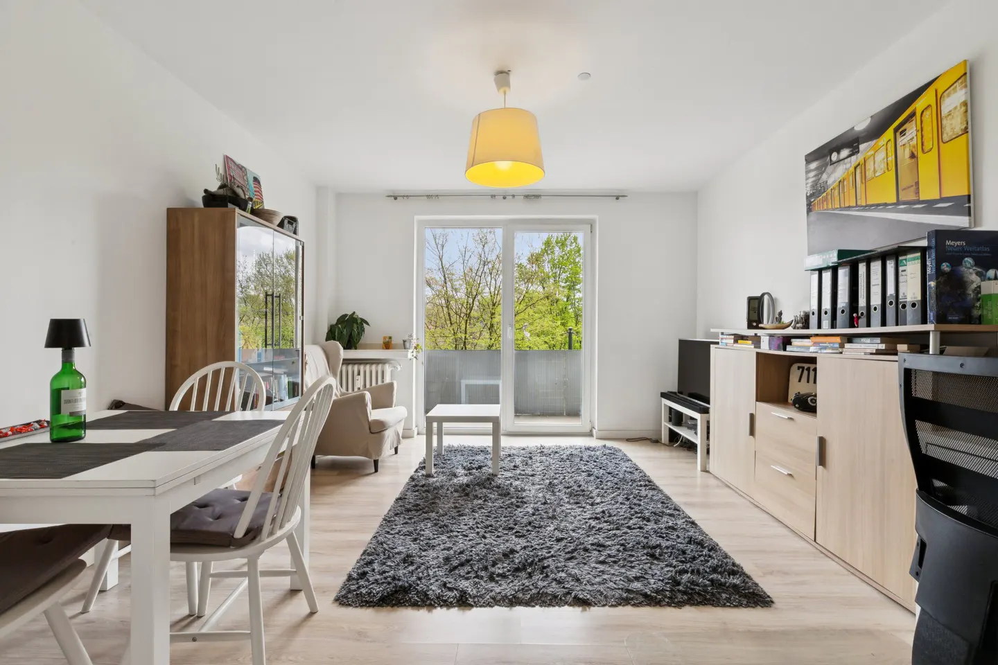 Bright living room with white walls, wood floors, and a gray rug. A dining table, armchair, and balcony are visible. A yellow subway art piece hangs on the wall.