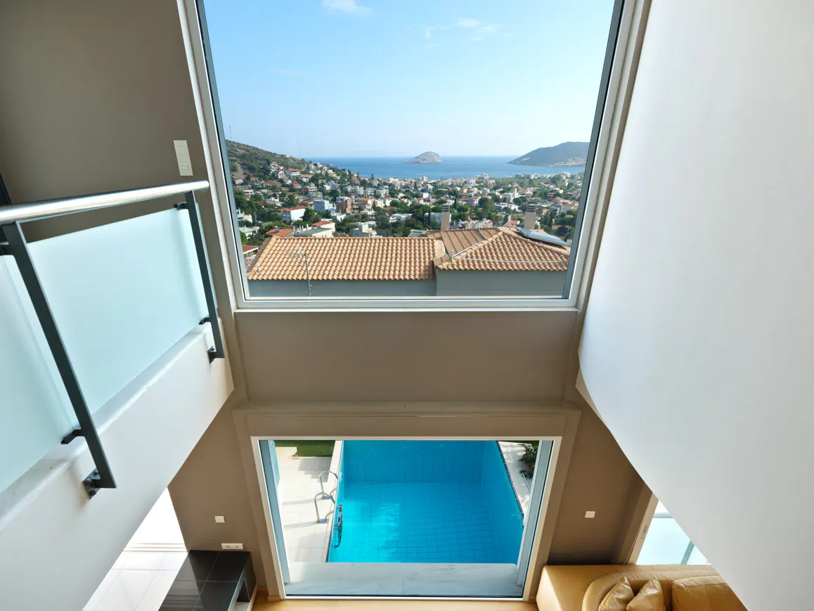 View from a modern interior looking down at a bright blue swimming pool and out to a coastal town and the sea.