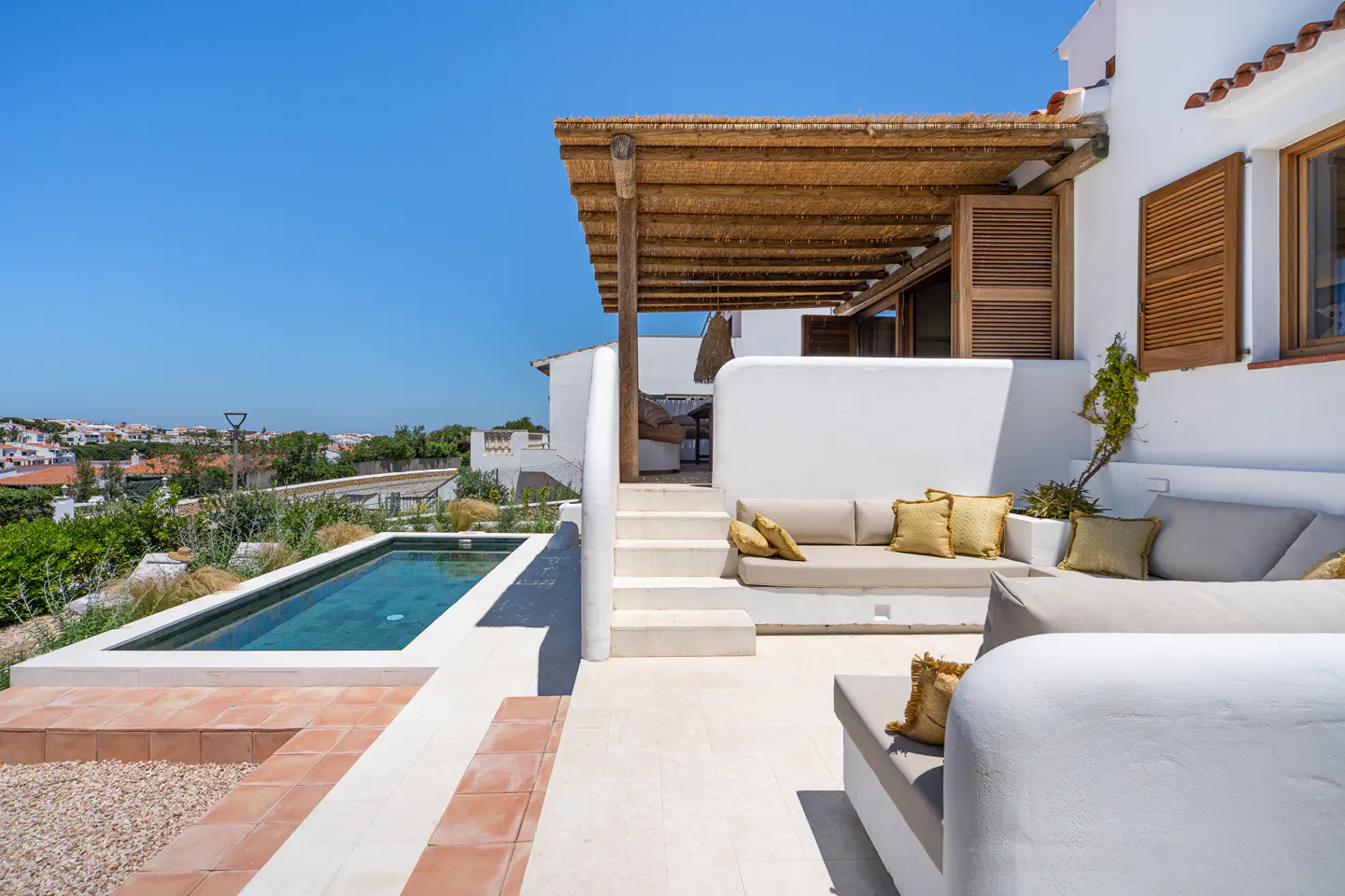 Outdoor patio with a pool, white walls, and beige cushions. A thatched roof covers part of the patio. Blue sky in the background.