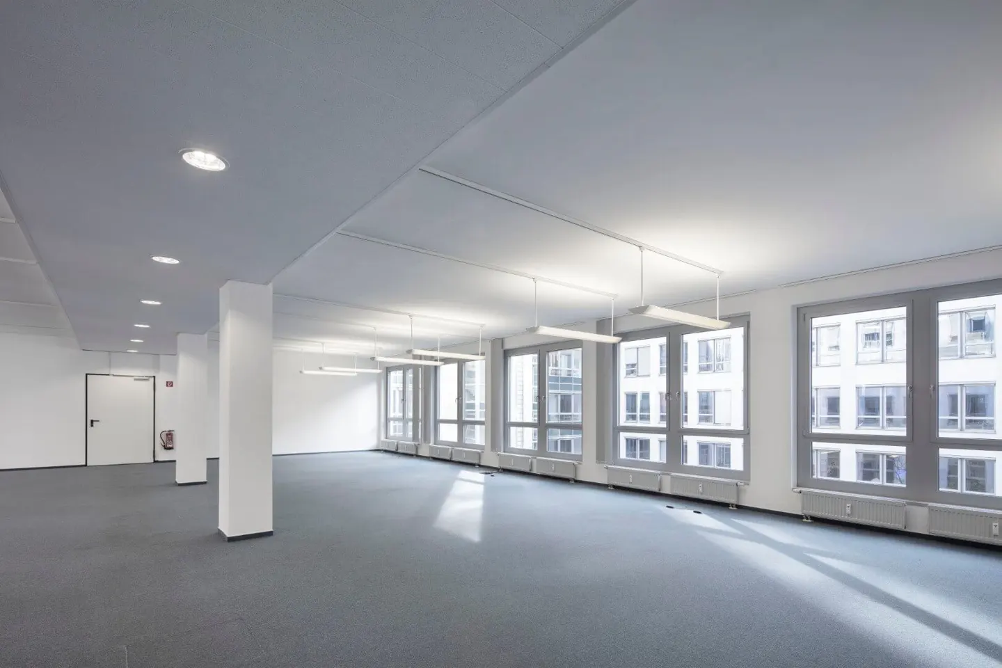 Bright, empty office space with gray carpet, white walls, and large windows. Columns and fluorescent lights are visible.