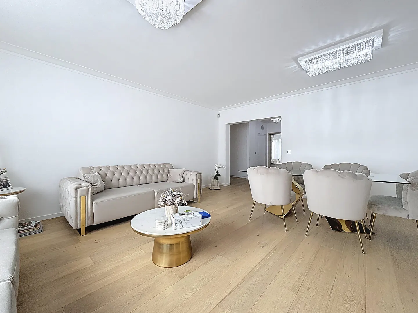 Bright living room with light wood floors, white walls, and elegant furniture. A beige tufted sofa and dining set with gold accents. Chandeliers on the ceiling.