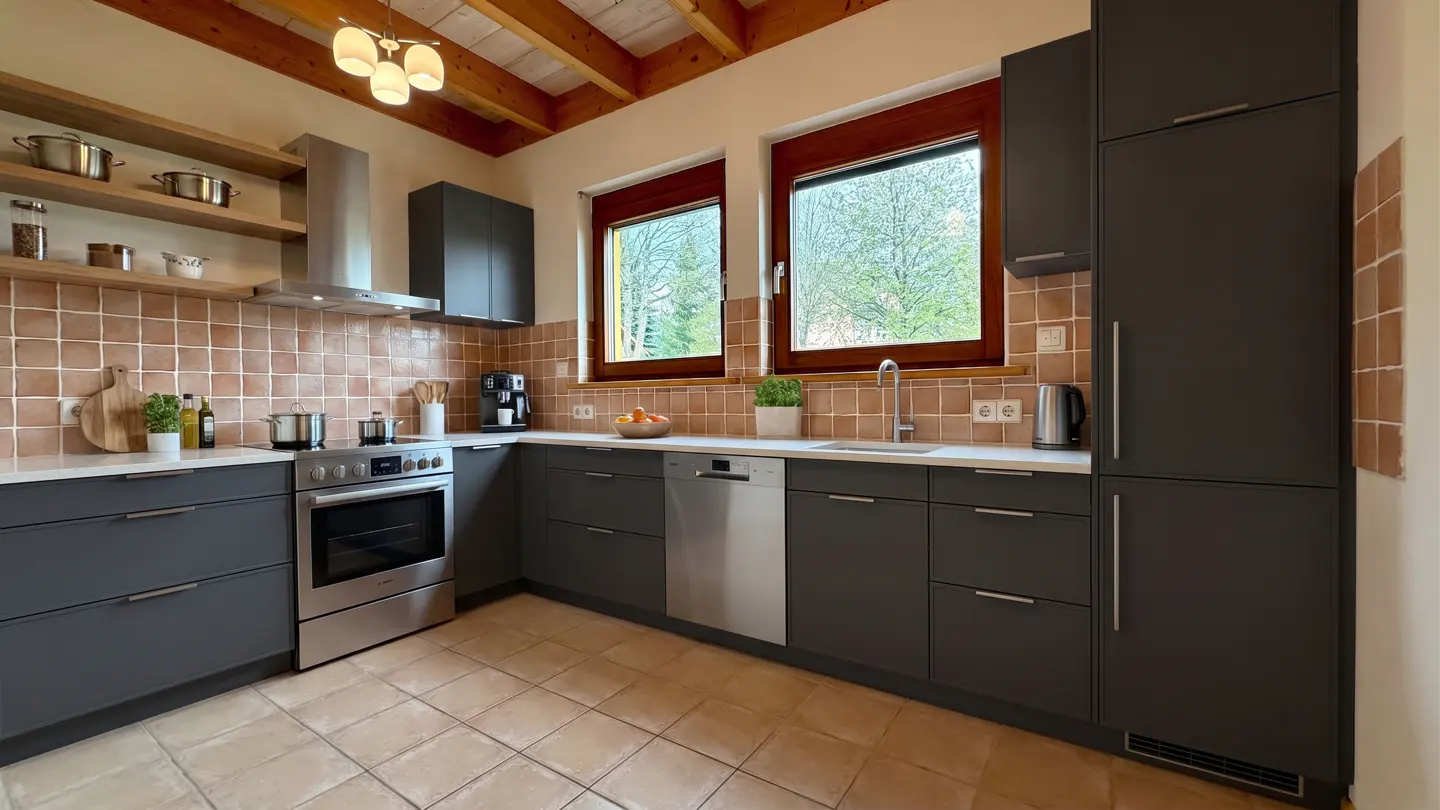 A modern kitchen with gray cabinets, stainless steel appliances, and terracotta tile backsplash and flooring. Two windows overlook a green landscape.