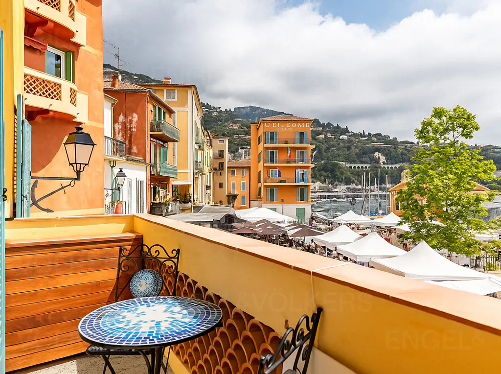 View from a balcony with a blue mosaic table overlooking a Mediterranean town square with colorful buildings and white tents.