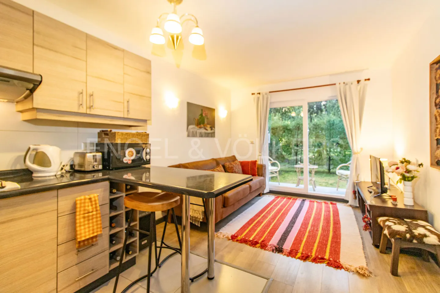 A bright apartment interior with a kitchen, brown sofa, and a view of a green garden through glass doors.