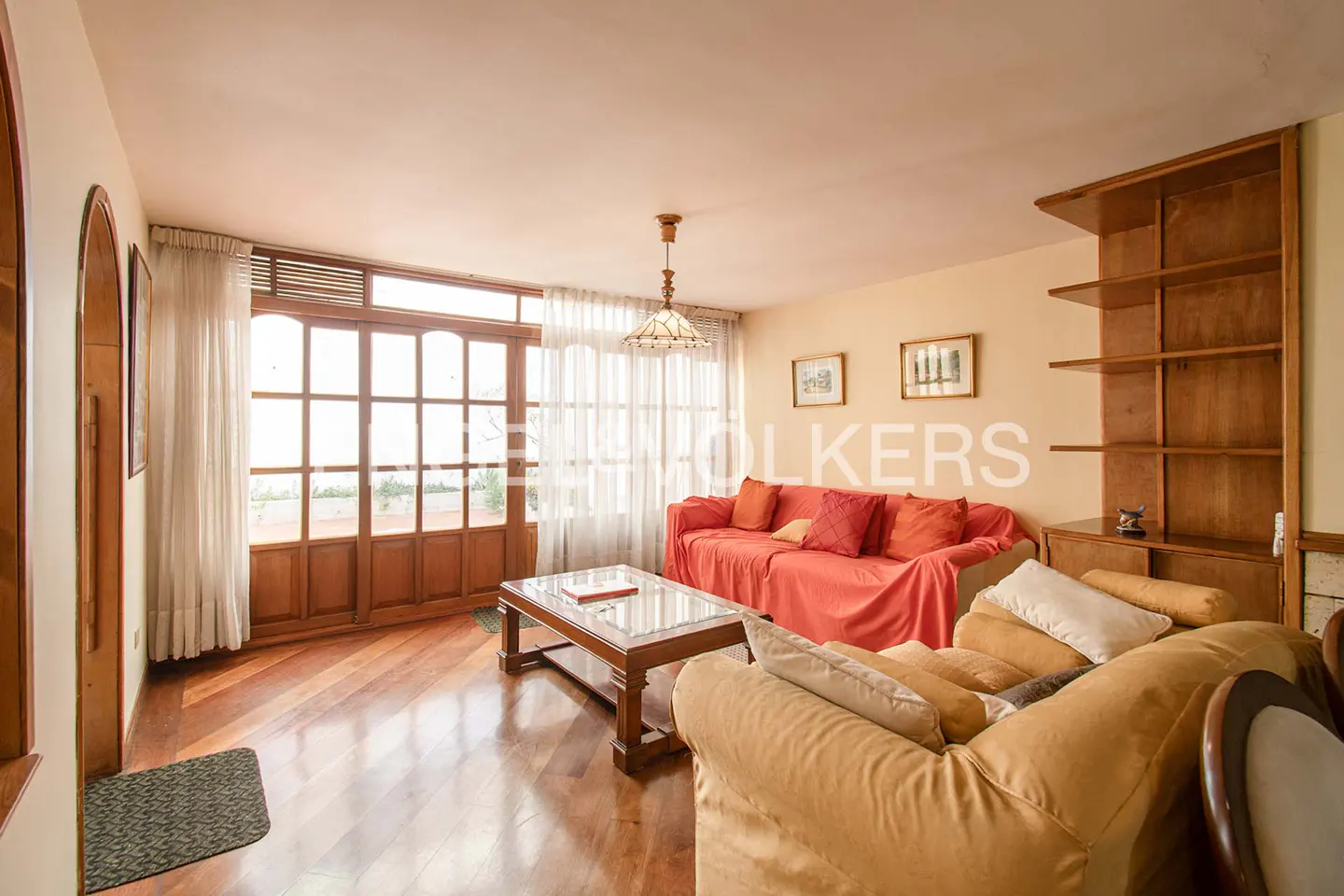 Living room with wood floors, a red sofa with pillows, and a tan armchair. A glass-topped coffee table sits in front of the sofa. A wooden bookshelf is on the right.