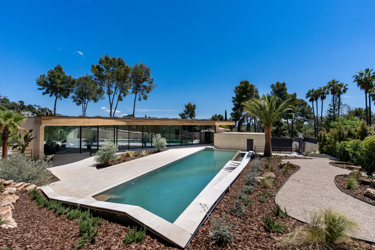 Modern home with a long, rectangular pool. The house has glass walls and a flat roof, surrounded by trees and landscaping. Blue sky above.