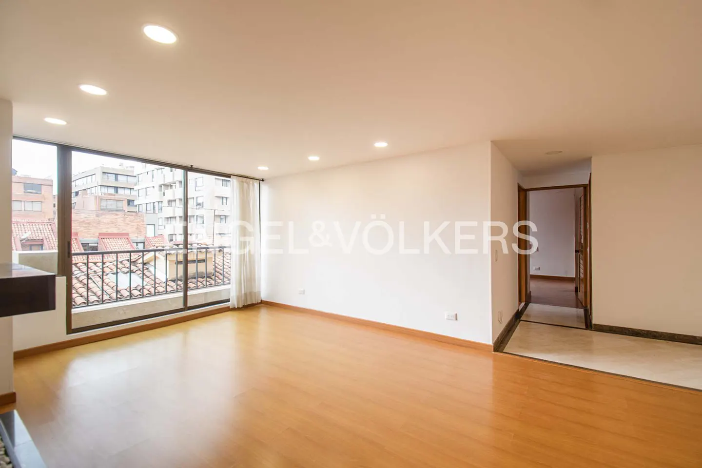 Bright, empty room with wood floors, white walls, and large windows overlooking a city. Doorway to another room is visible. Engel & Völkers logo.