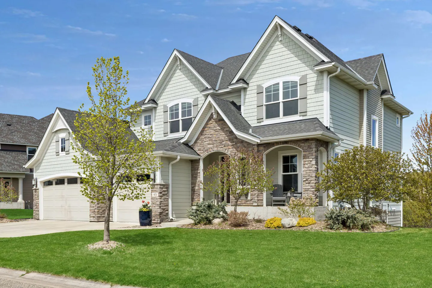 Two-story house with light green siding, stone accents, gray shutters, and a green lawn under a blue sky.