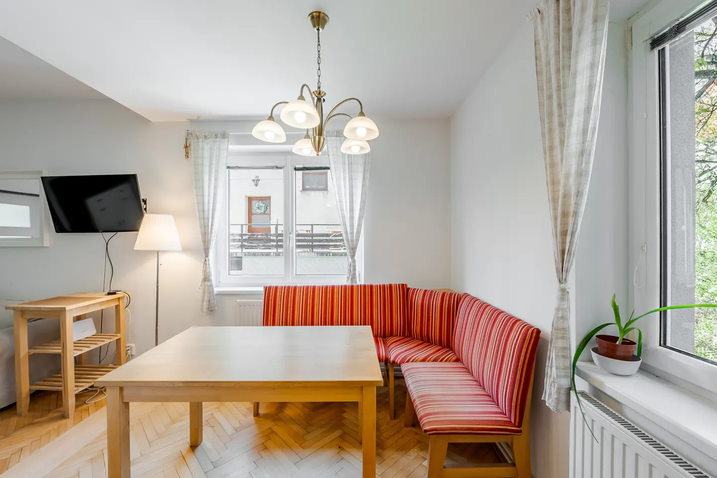 Bright dining area with a wooden table, striped corner bench, and chandelier. Windows with sheer curtains let in natural light. TV on the wall.