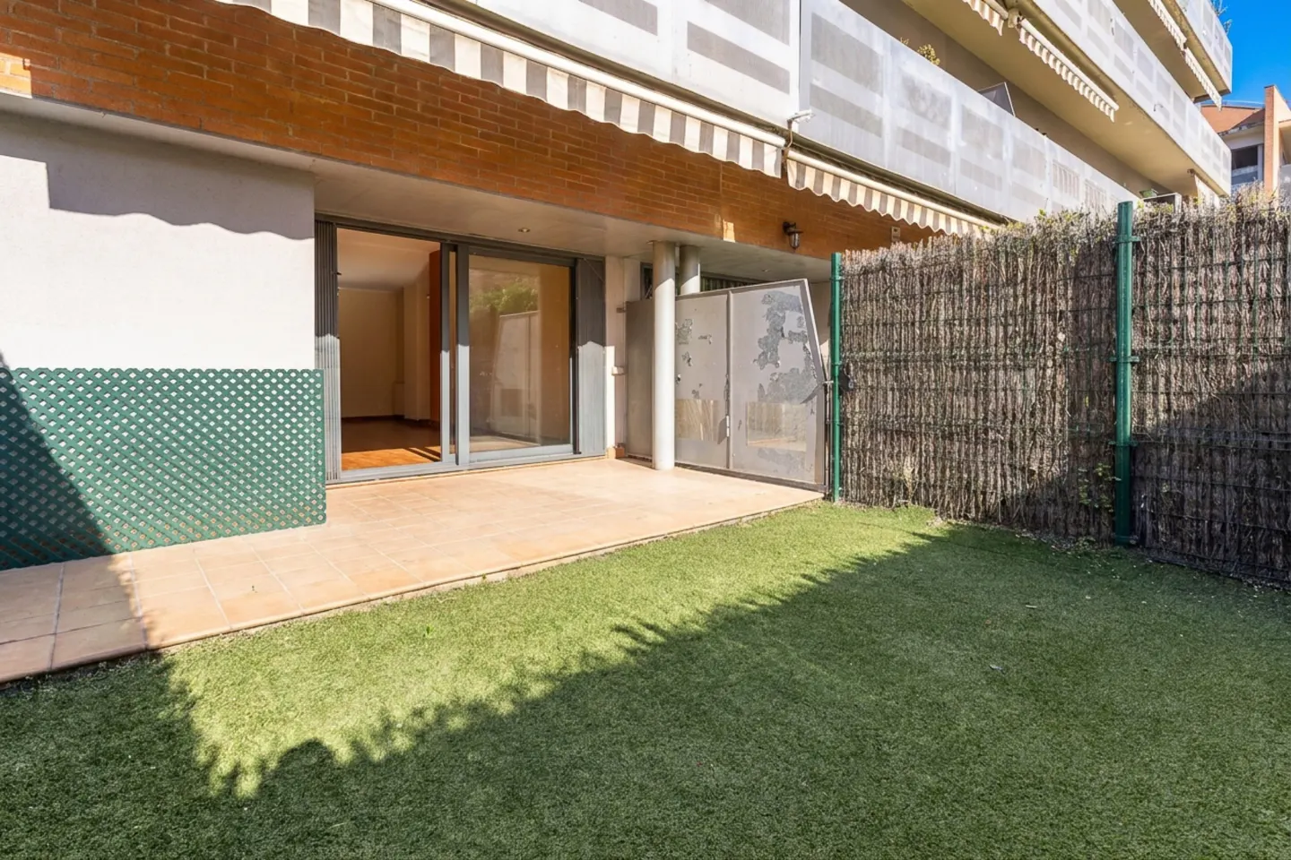 A ground-level patio with sliding glass doors opens to a small green lawn enclosed by a woven fence.