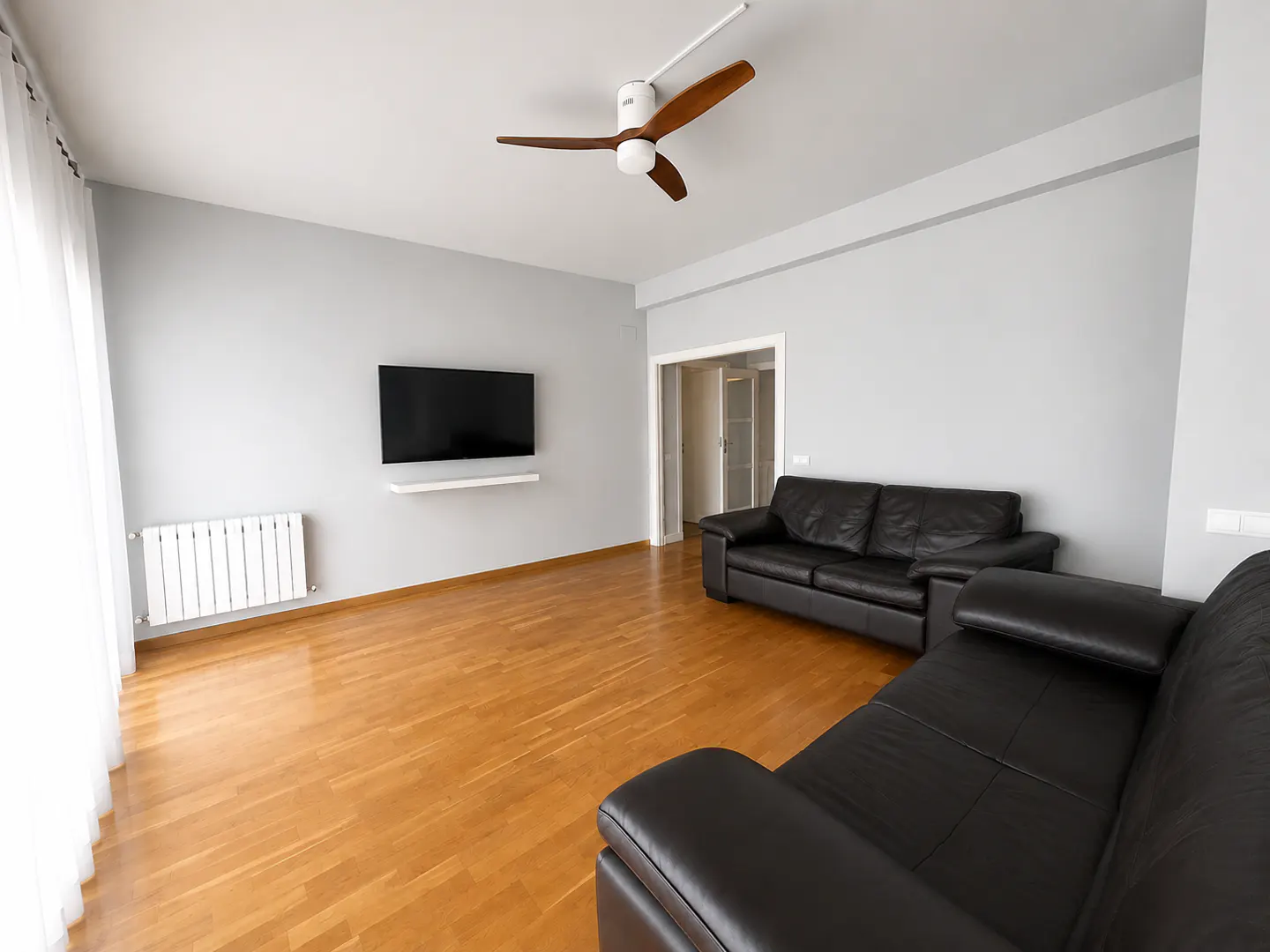 Living room with light gray walls, hardwood floors, two black leather sofas, a wall-mounted TV, and a wooden ceiling fan.